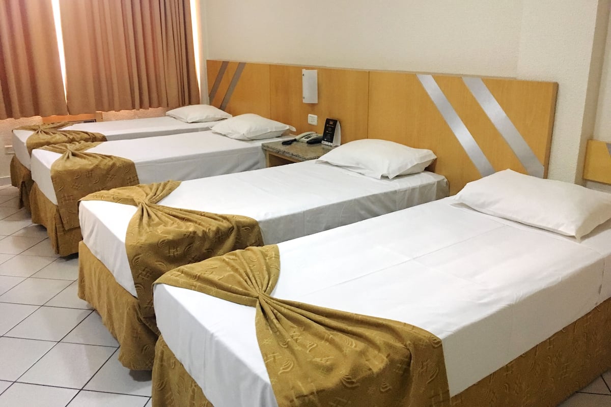 A hotel room with three single beds, each with a white mattress and a brown bed skirt. The room has a wooden headboard with a diagonal stripe design, and there is a nightstand with a phone and a small table beside the beds.