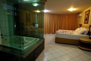 Habitación de hotel con un jacuzzi de vidrio, una cama con ropa blanca y una mesita de madera.