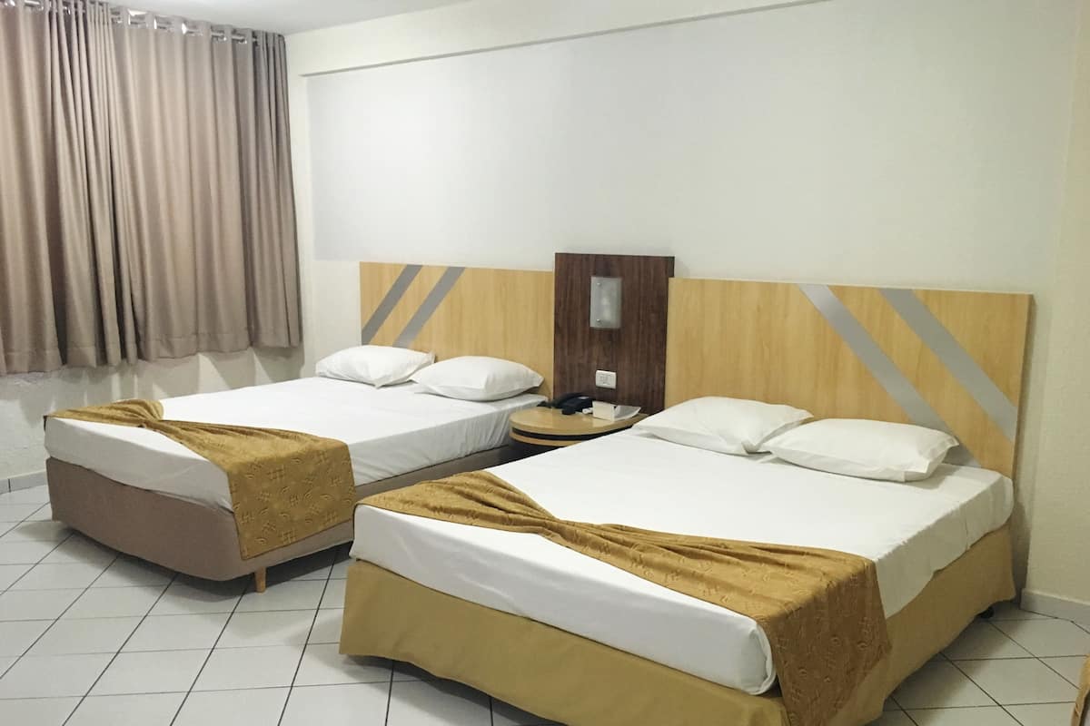 A hotel room with two beds, each with a brown bedspread and white pillows. There is a wooden headboard with a diagonal stripe design, a nightstand with a lamp, and a window with beige curtains.