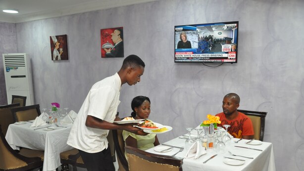 A dining area with tables set for two, a television mounted on the wall displaying a news broadcast, and artwork on the walls. - Grand Capital Hotel (Akure)