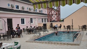 An outdoor swimming pool area with lounge chairs and tables, surrounded by a building with a pink facade and a red metal structure. - Grand Capital Hotel (Akure)
