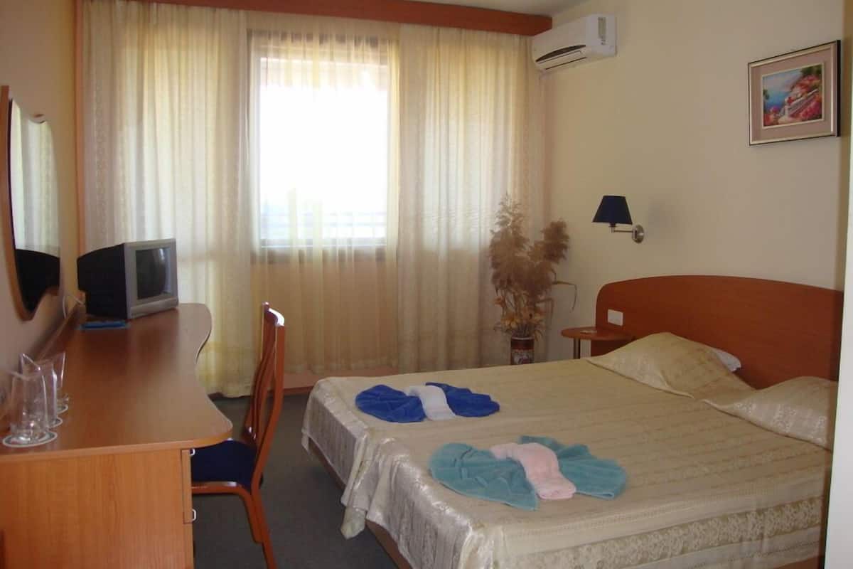 Quarto de hotel com cama, escrivaninha com computador, cadeira, luminária e janela com cortinas.