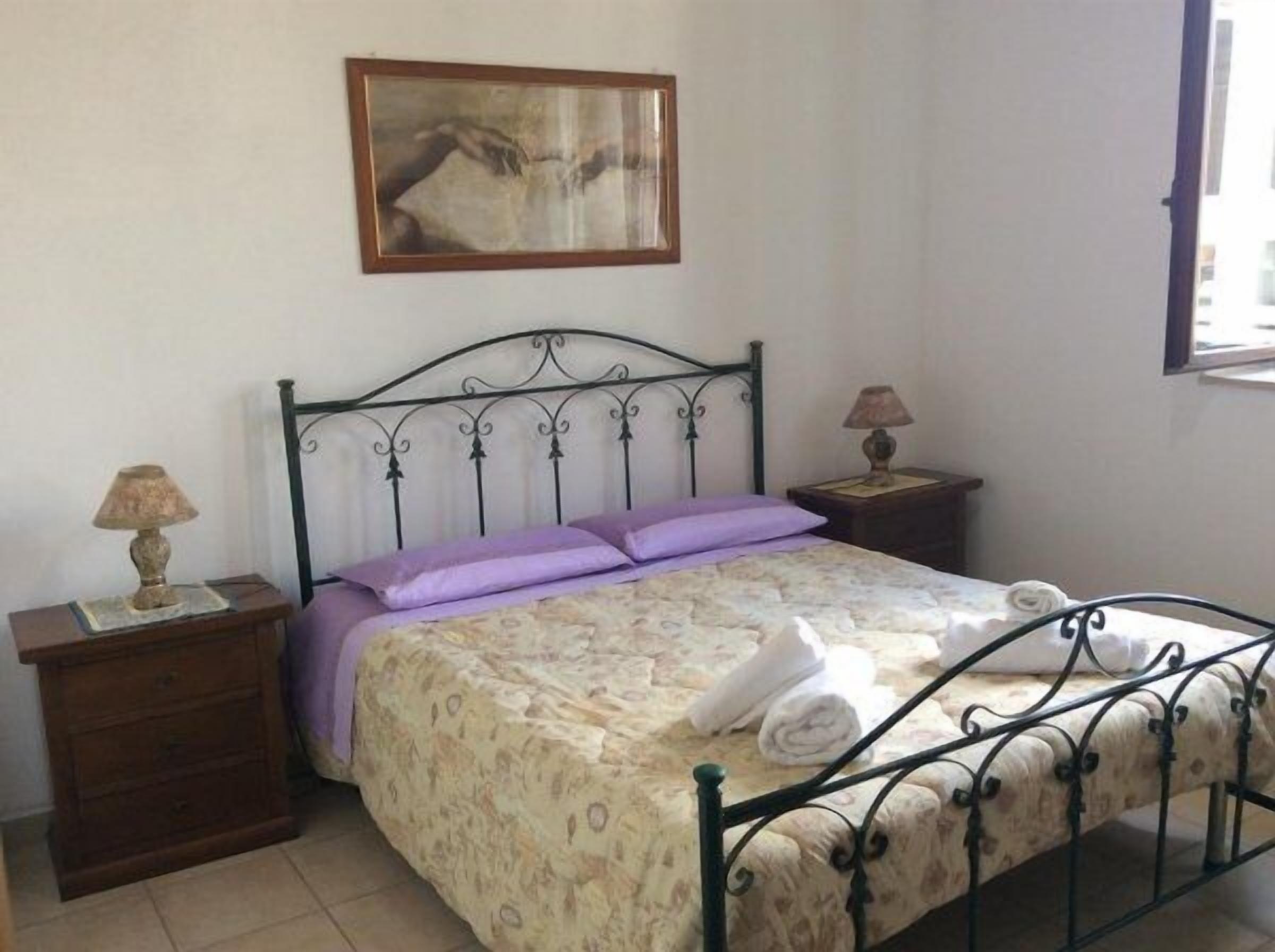 Ein Schlafzimmer mit einem Bett, zwei Nachttischen, einer Lampe auf jedem Tisch sowie einem Gemälde über dem Bett und einem Fenster rechts.