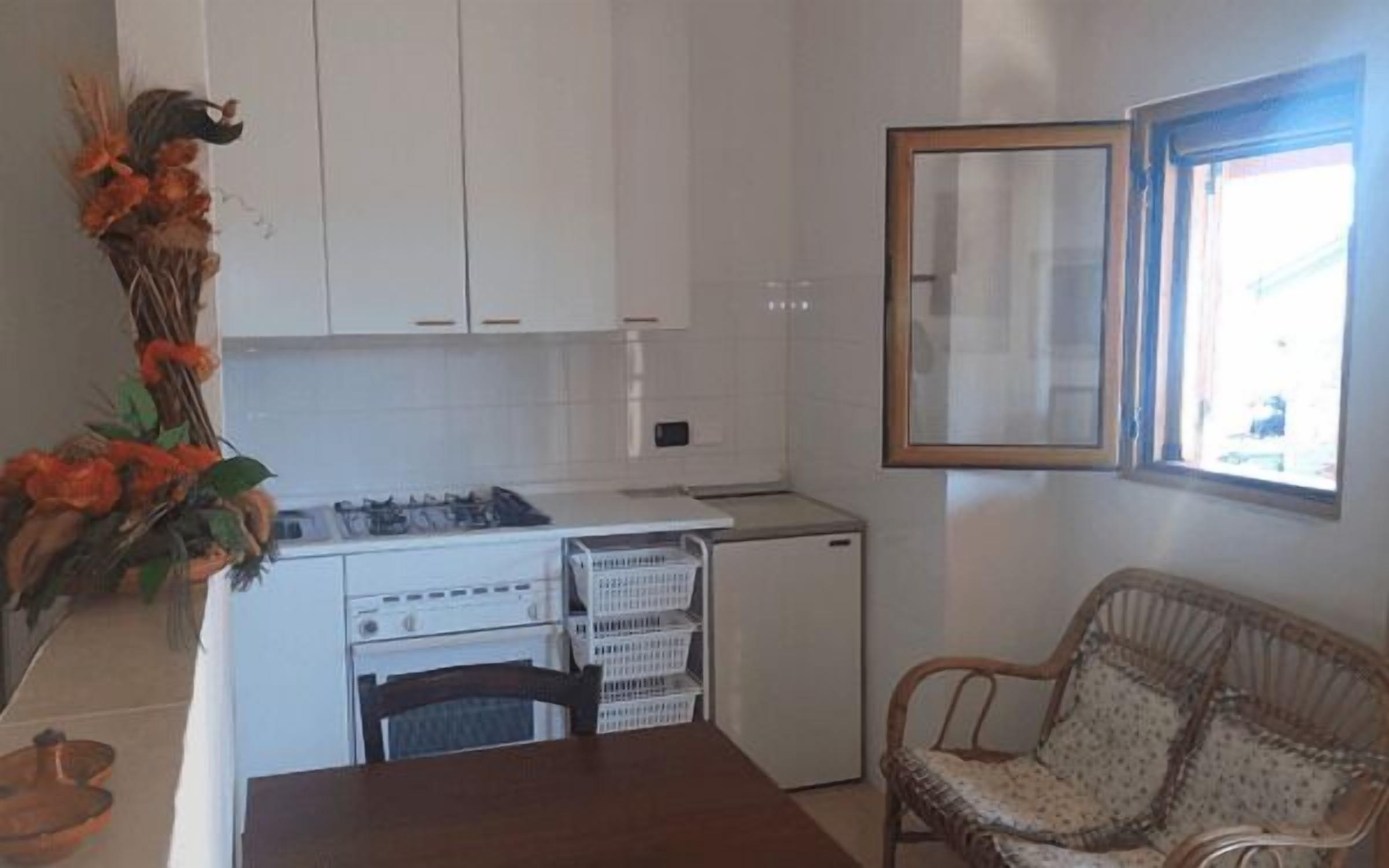 A compact kitchen with white cabinets, a stove, and a small refrigerator. There is a dining table with a chair and a wicker sofa. A window with a wooden frame is visible.