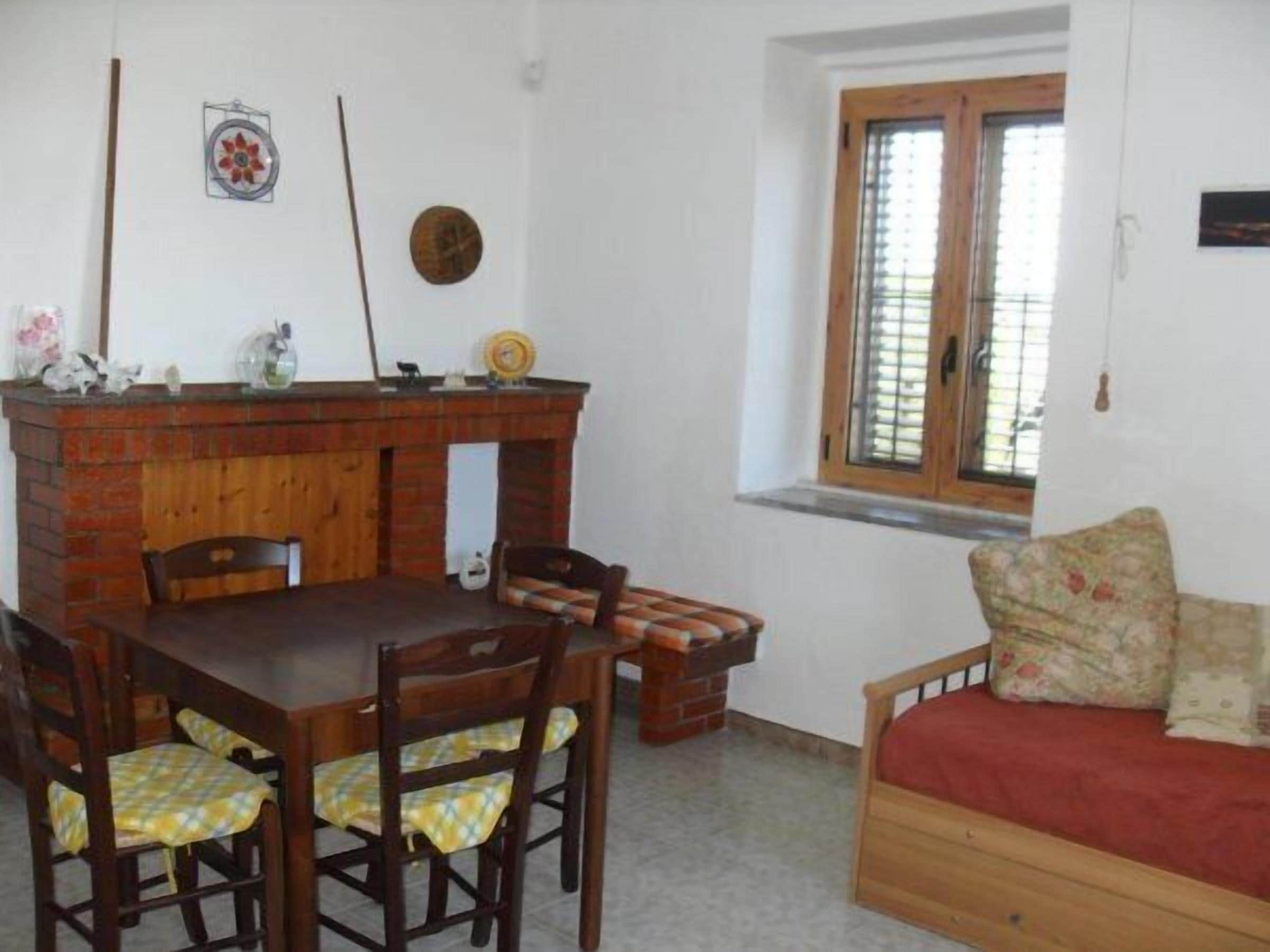 Um quarto com uma mesa de jantar de madeira, cadeiras, um sofá, uma lareira e uma janela com persianas.