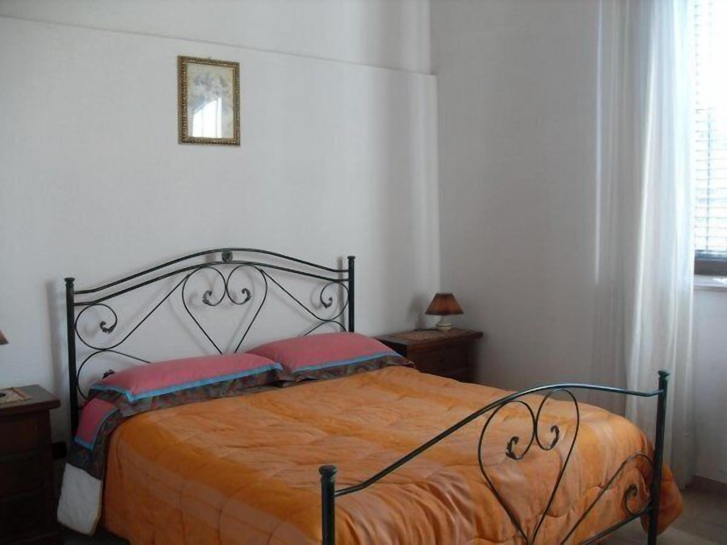 A bedroom with a metal bed frame, an orange bedspread, two pillows, a bedside table with a lamp, and a window with curtains.