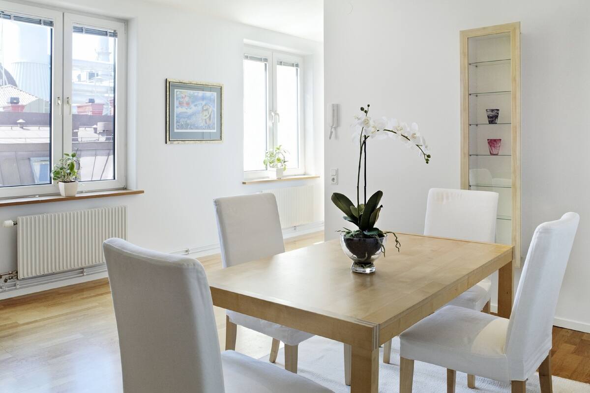 A dining area with a wooden table, four white chairs, a potted orchid, and a framed picture on the wall.