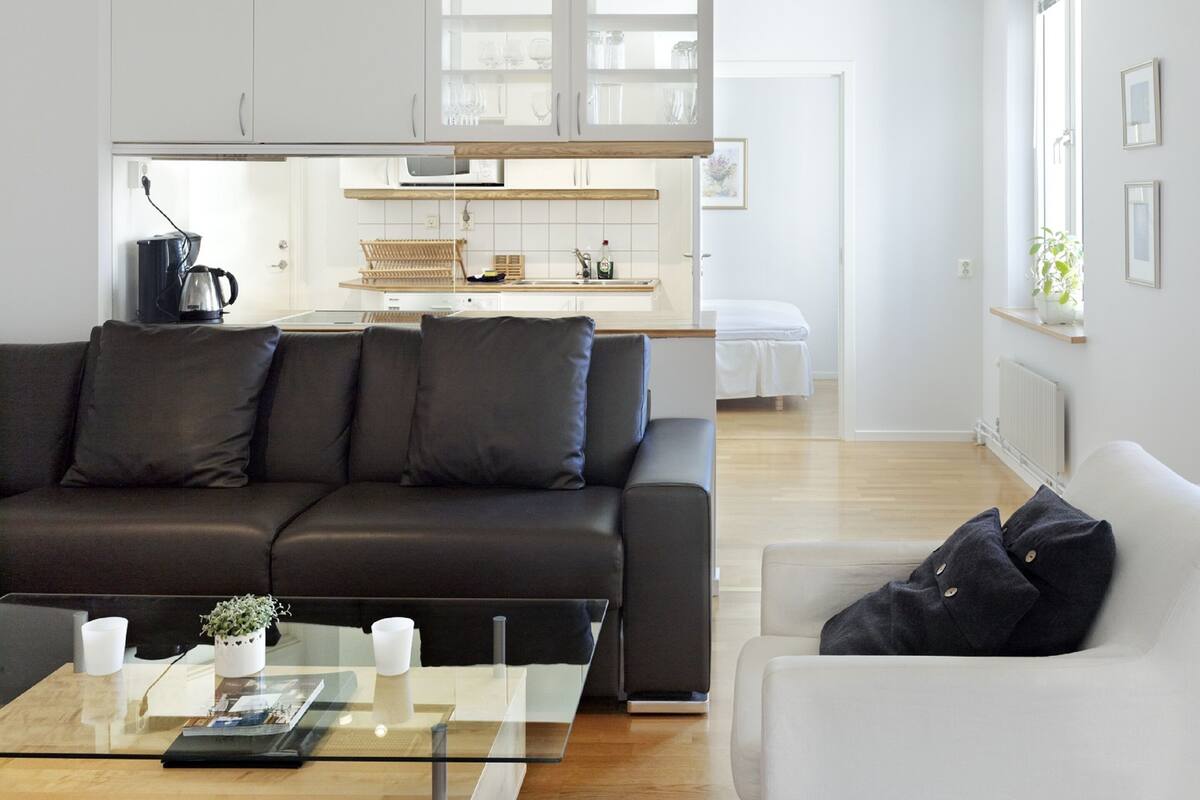A modern living room with a black leather sofa, a glass coffee table, and a white armchair. A kitchen with white cabinets and a wooden countertop is visible in the background.
