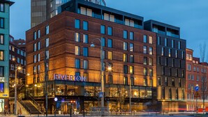 Un hotel de varios pisos con un llamativo letrero 'RIVERTON', que cuenta con una fachada moderna de ladrillo, ventanas grandes y una entrada visible con una escalera.