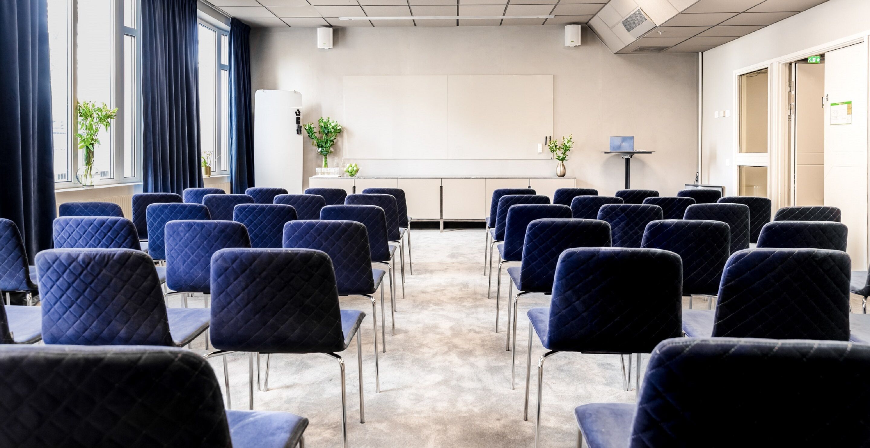 A conference room with rows of blue chairs, a whiteboard, and two potted plants.