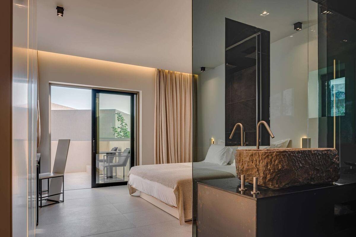 A modern hotel room with a bed, a glass shower enclosure, a wooden vanity, and a balcony with a table and chairs.