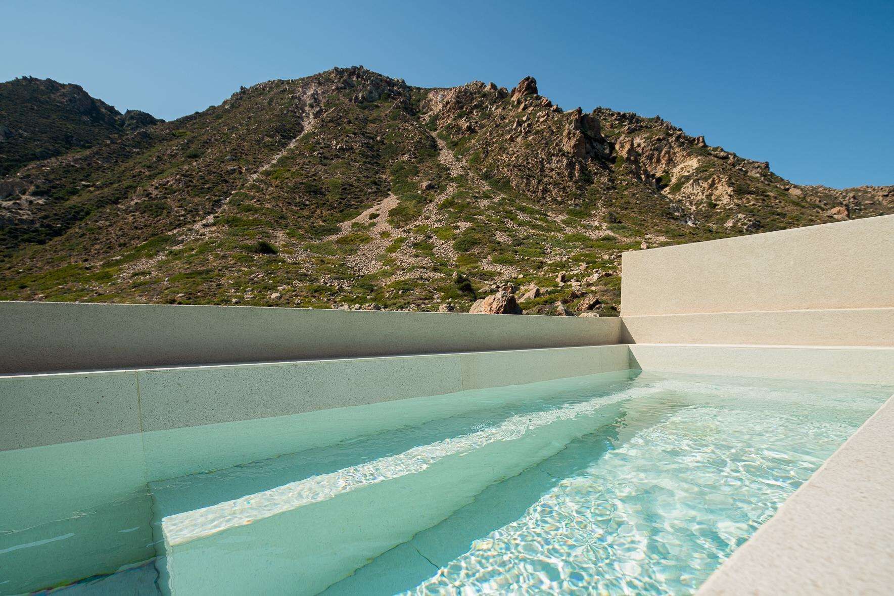 Una piscina con agua cristalina y escalones de acceso, rodeada de terreno rocoso y montañas.