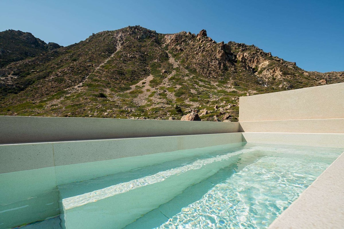 A swimming pool with clear water and steps leading into it, surrounded by rocky terrain and mountains.