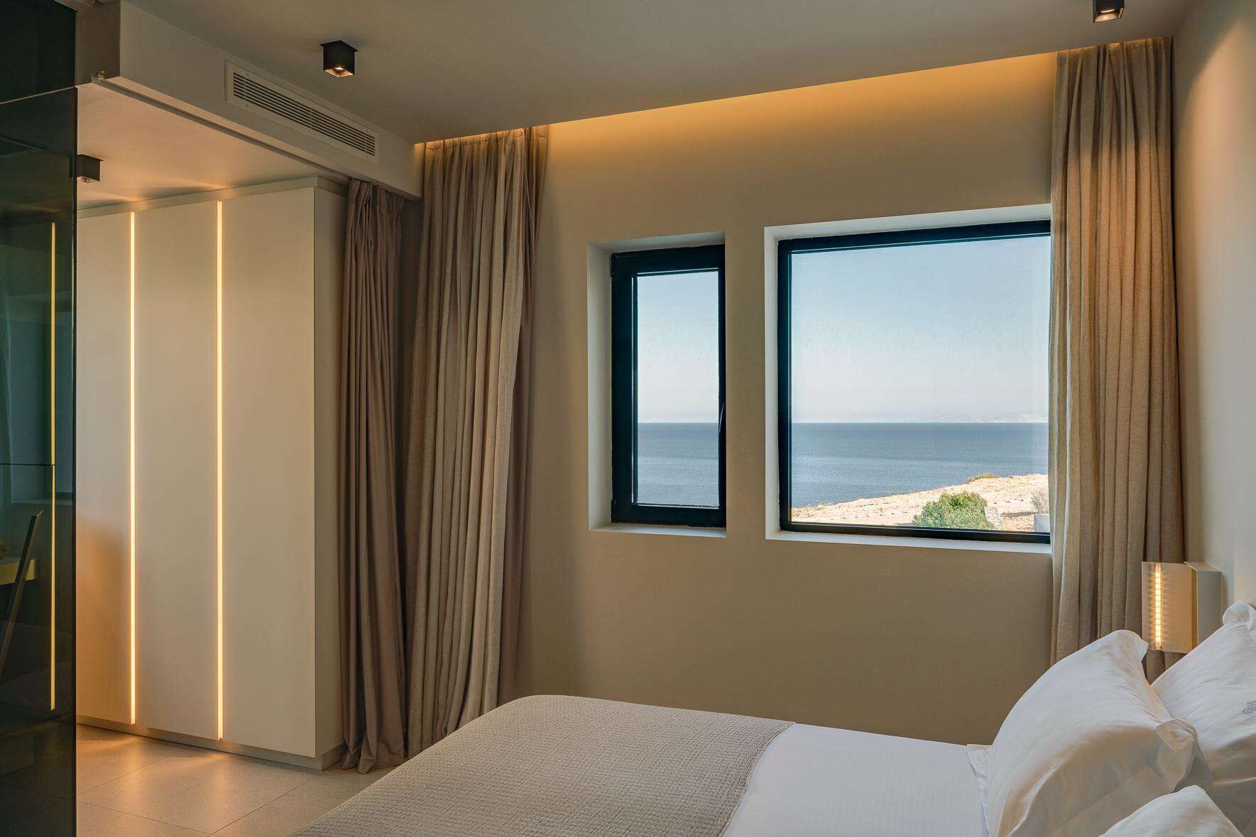 A bedroom with a bed, two windows with a view of the sea, and a bedside lamp.