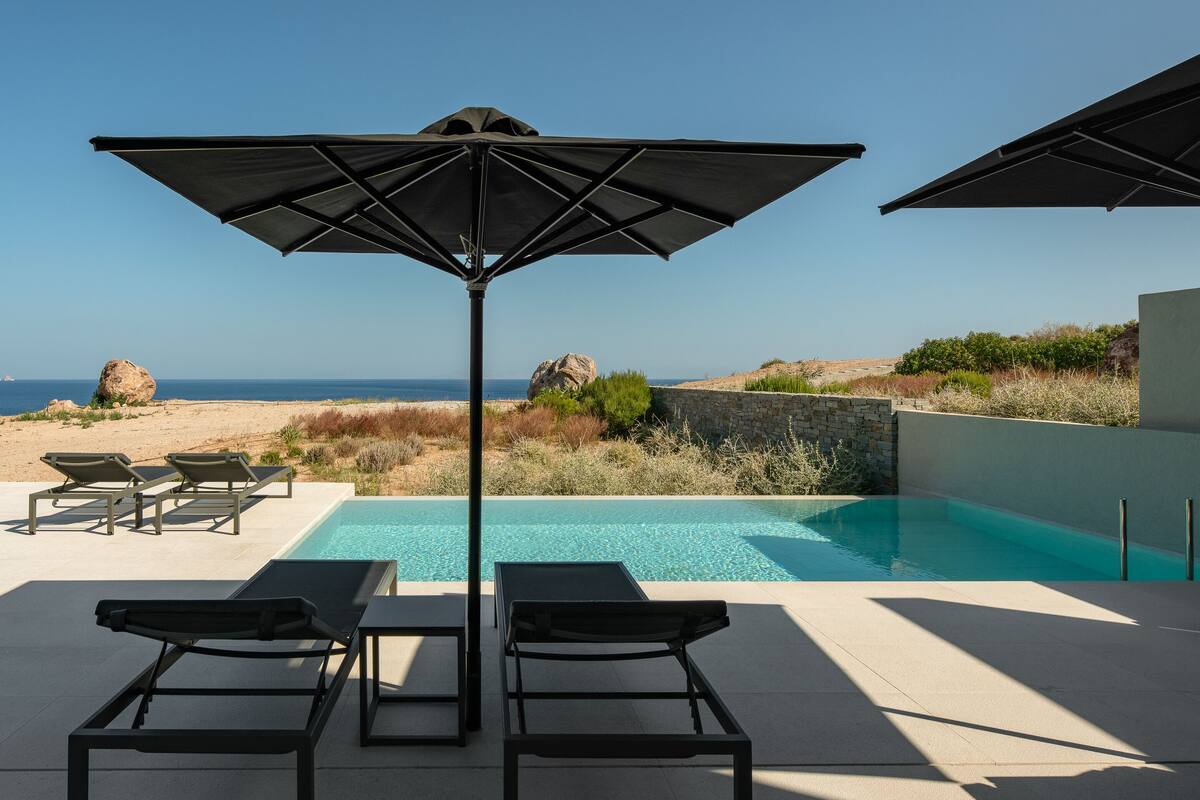 A poolside area with lounge chairs and umbrellas, overlooking a clear blue sea and rocky landscape.