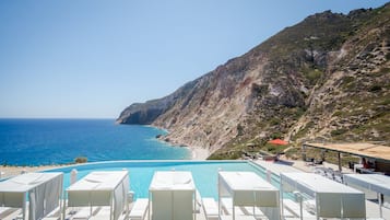 Piscina con sillones blancos, una sombrilla roja y vistas al mar y una ladera rocosa.