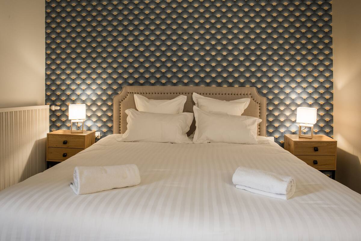 Un lit bien fait, avec du linge de lit blanc et des oreillers, deux tables de chevet avec des lampes et un mur à motifs derrière le lit.