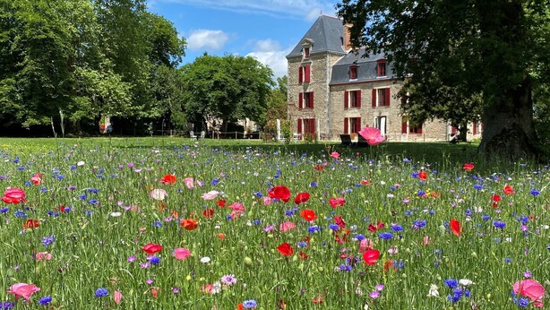 Un grand bâtiment en pierre, doté de volets rouges et d’un jardin où poussent de nombreuses variétés de fleurs.