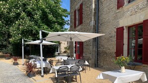 Une terrasse extérieure avec des tables et des chaises, un parasol blanc et, en arrière-plan, un bâtiment en pierre avec des volets rouges.
