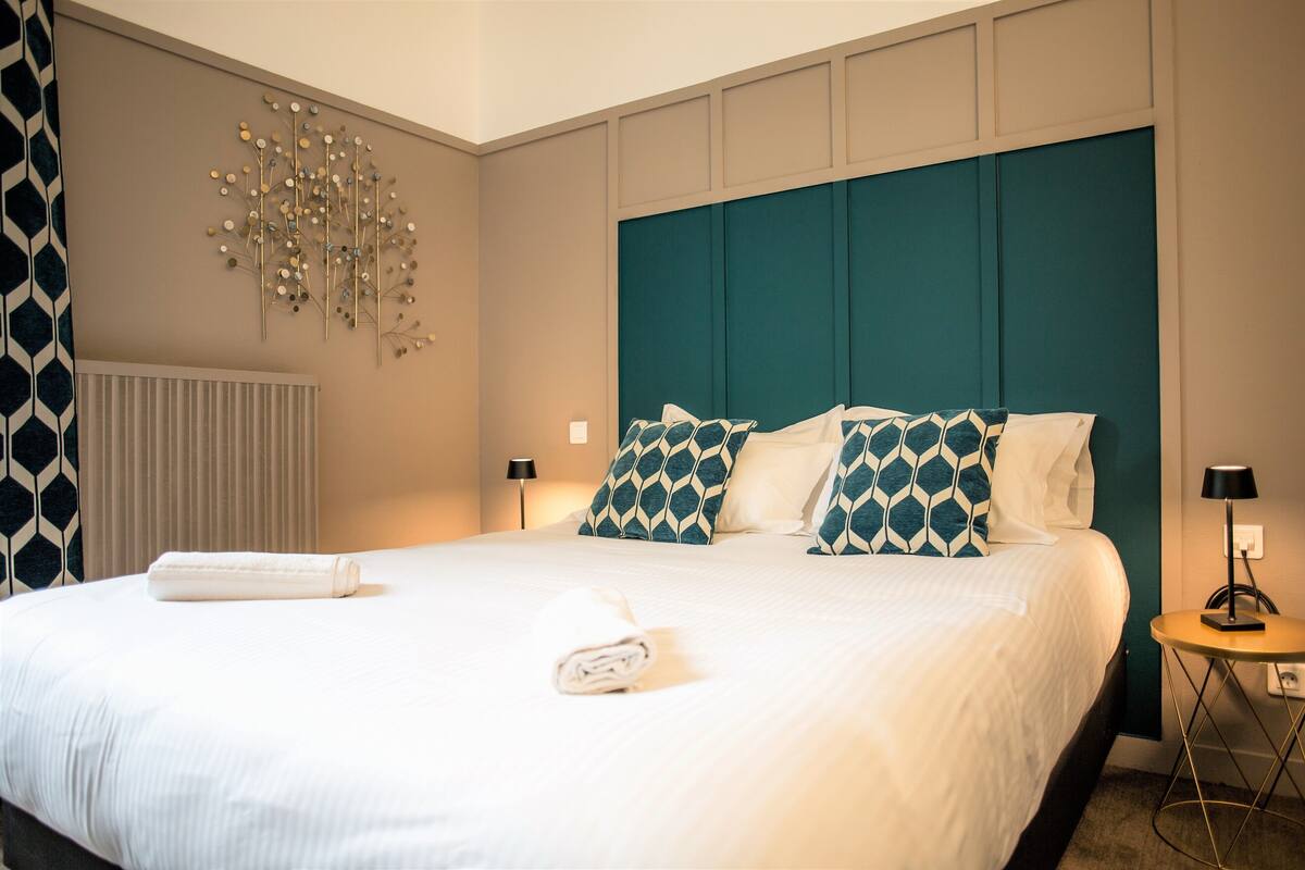 Un lit bien fait, avec du linge de lit blanc et des coussins à motifs turquoise, un luminaire fixé au mur et une petite table de chevet avec une lampe.
