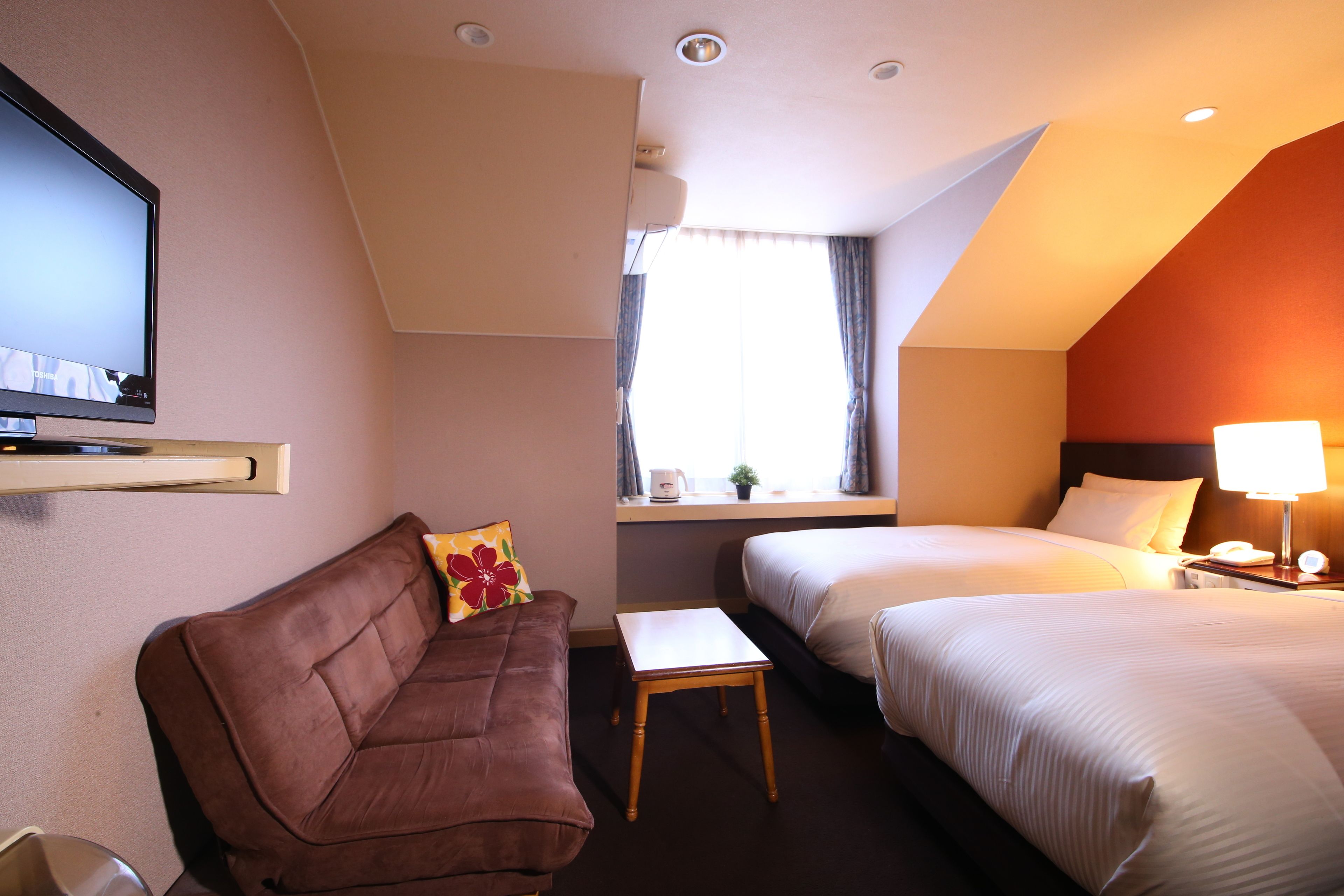 A hotel room with a brown sofa, a small table, two beds, a TV, a window with curtains, and a lamp on the nightstand.