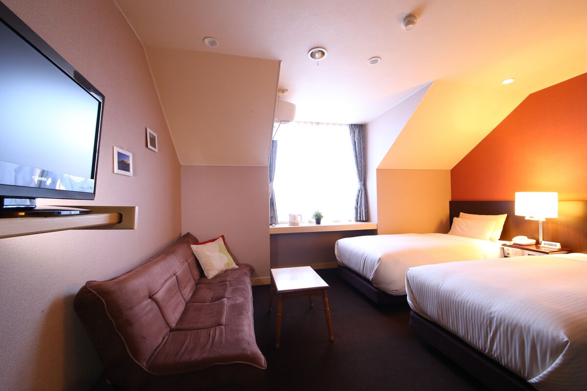 A hotel room with a sofa, two beds, a TV, a small table, and a window with curtains.