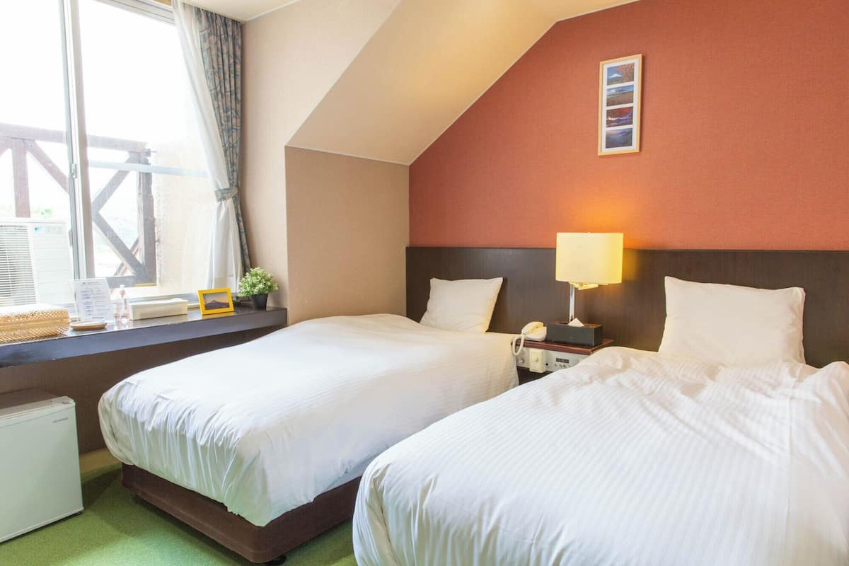 A hotel room with two beds, a nightstand with a lamp, a window with curtains, and a view of the outside.