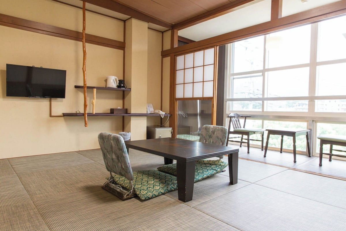 A traditional Japanese-style room with tatami flooring, a low table, and chairs. A television is mounted on the wall, and there are sliding doors leading to a balcony.