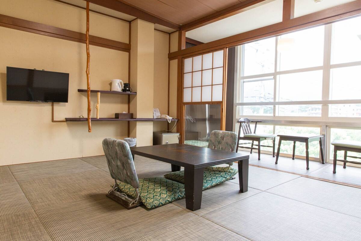 A traditional Japanese-style room with tatami flooring, a low table, chairs, a television, and a large window.