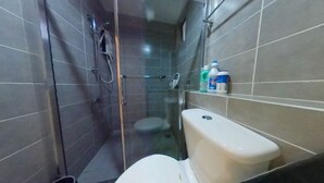 A bathroom with a toilet, a glass-enclosed shower, and a shelf with cleaning products. - TnJ Home (Kota Kinabalu)