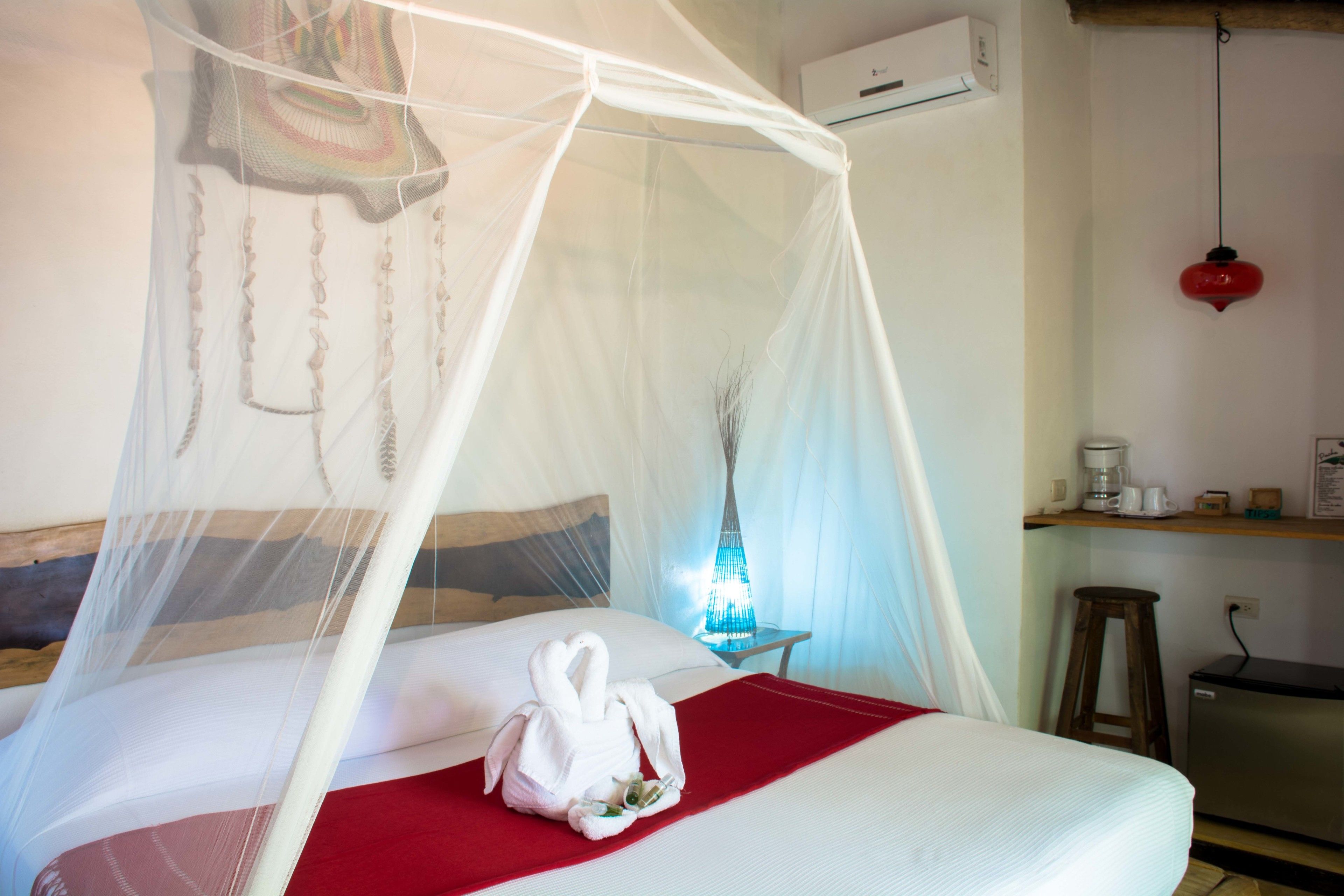 Habitación de hotel con cama, manta roja, toalla blanca, mosquitero, mesita de noche con lámpara, cafetera y refrigerador pequeño.