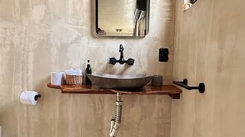 Un baño con un lavamanos grande, un espejo, un perchero para toallas y un estante de madera con artículos de aseo.