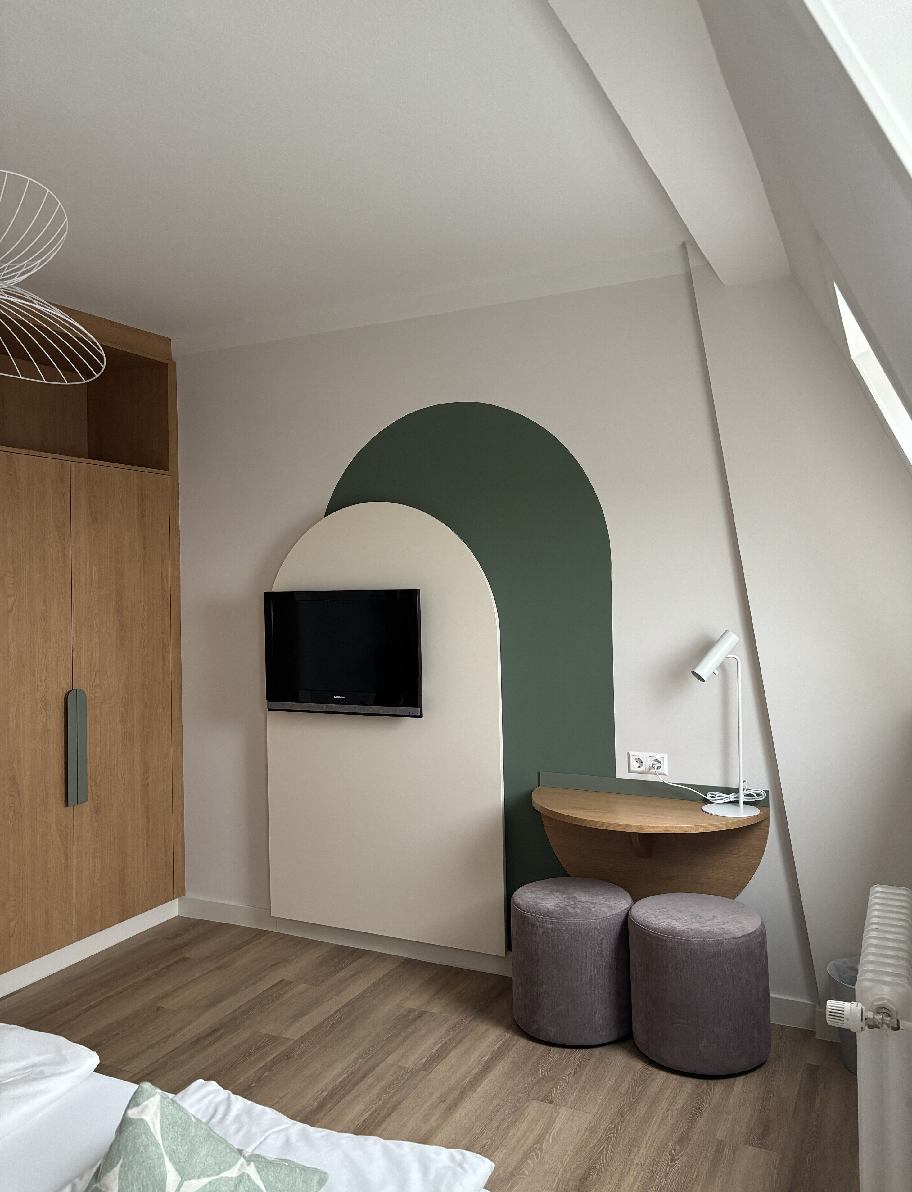 A modern bedroom with a wooden wardrobe, a mounted TV, a desk with a lamp, and two ottomans.