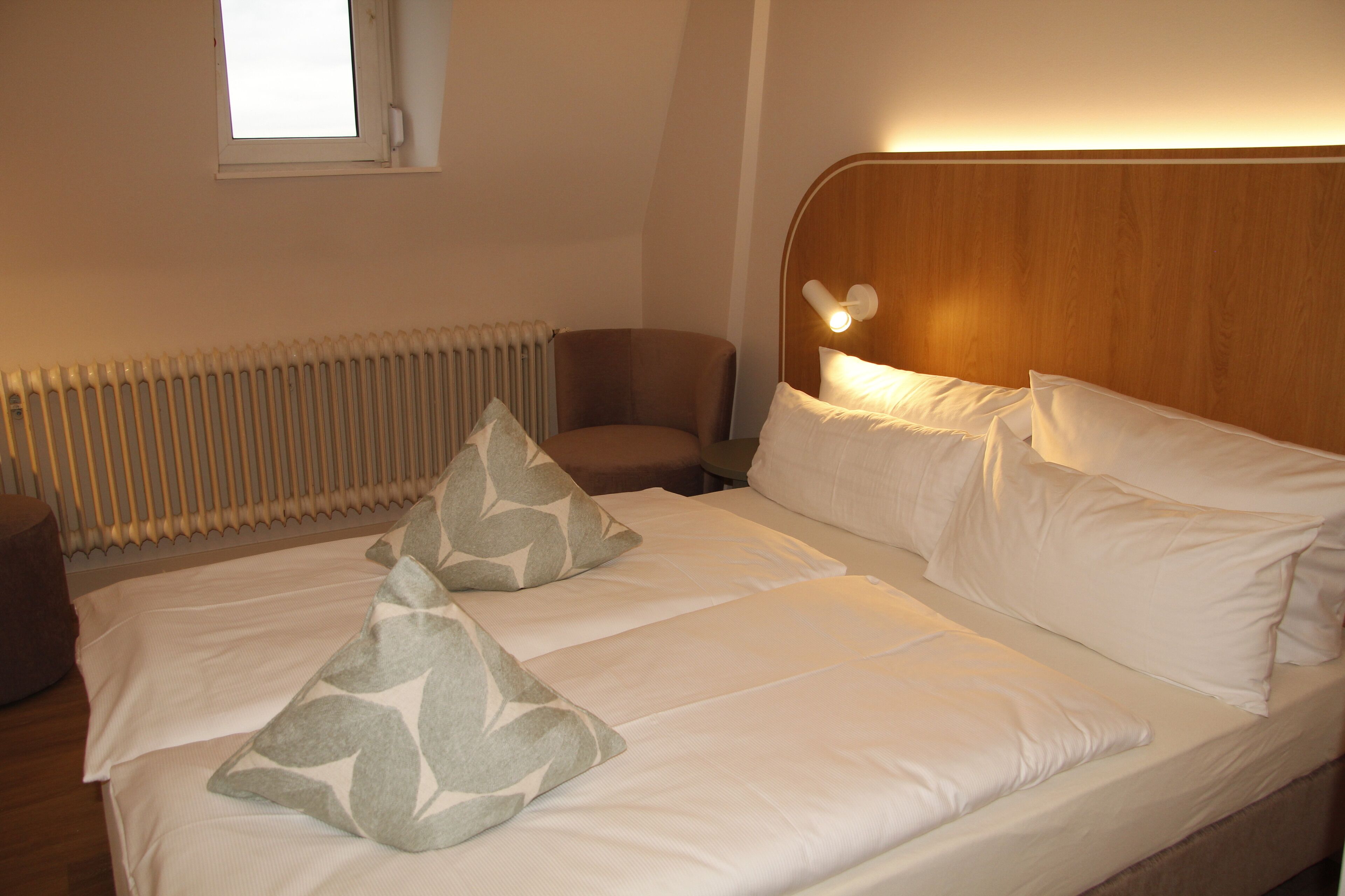 Habitación de hotel con cama, dos almohadas, una silla y un radiador.