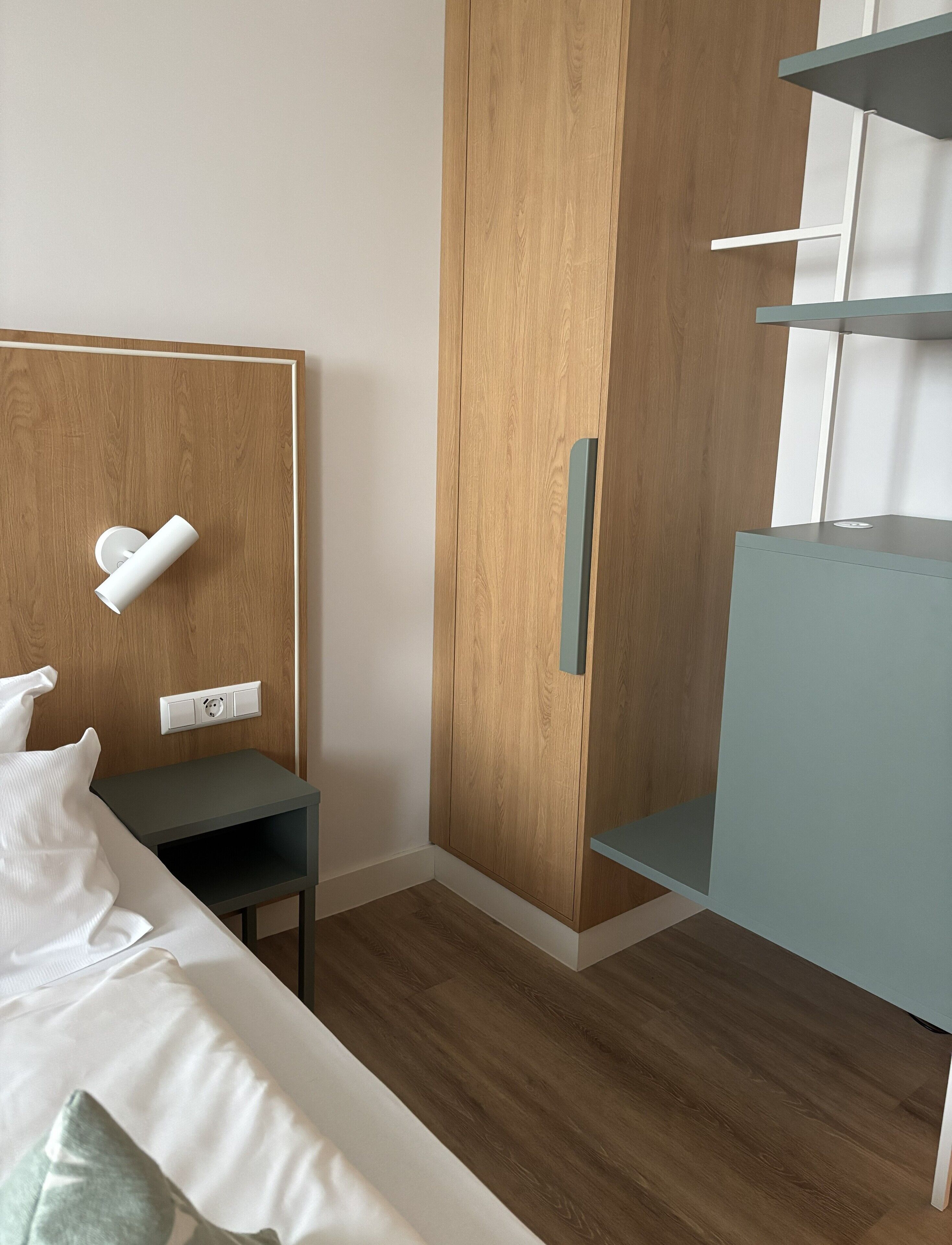 A bedroom with a wooden wardrobe, a bed with white bedding, a small bedside table, and a blue cabinet.