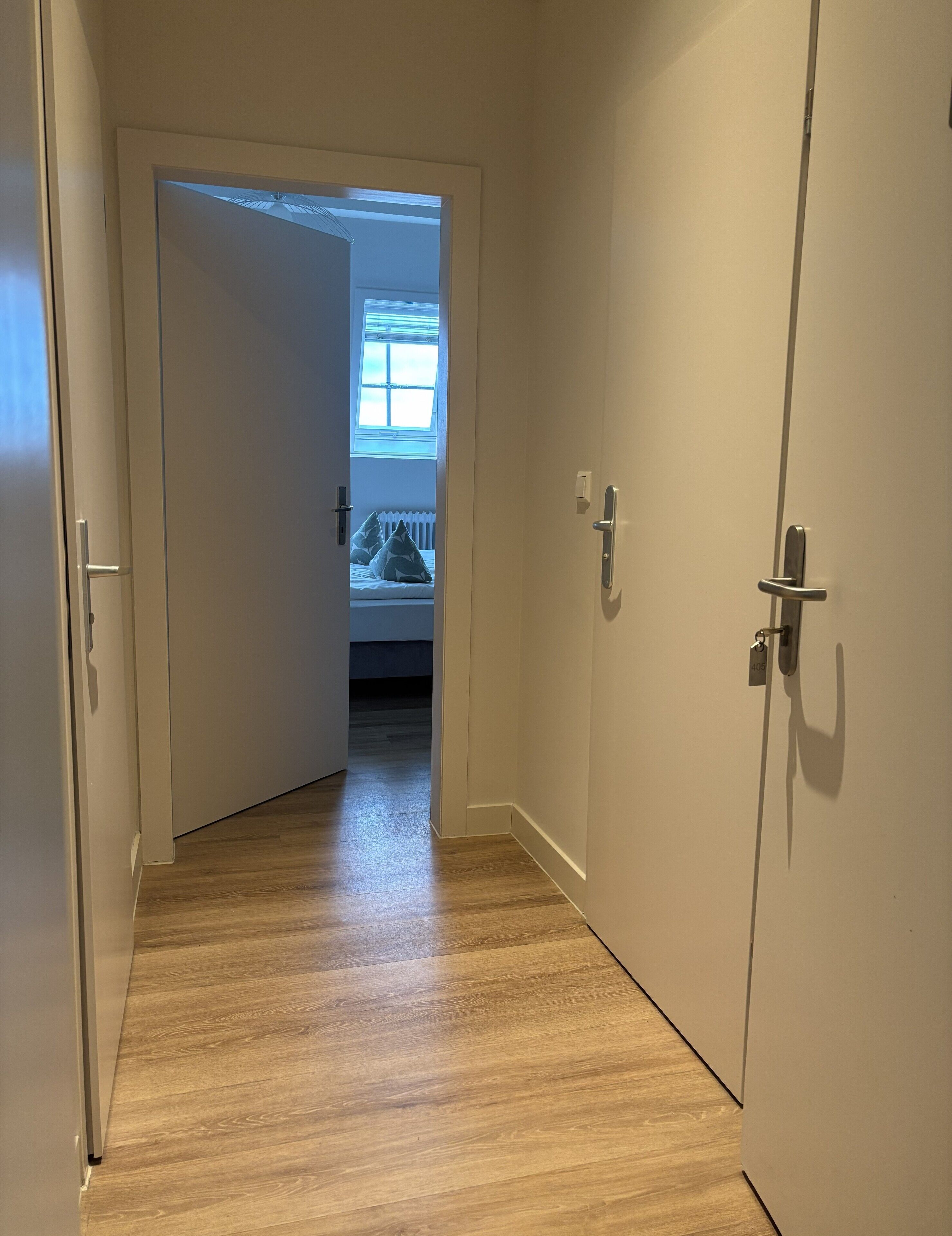 Un pasillo estrecho con suelo de madera que lleva a un dormitorio con ventana y radiador.
