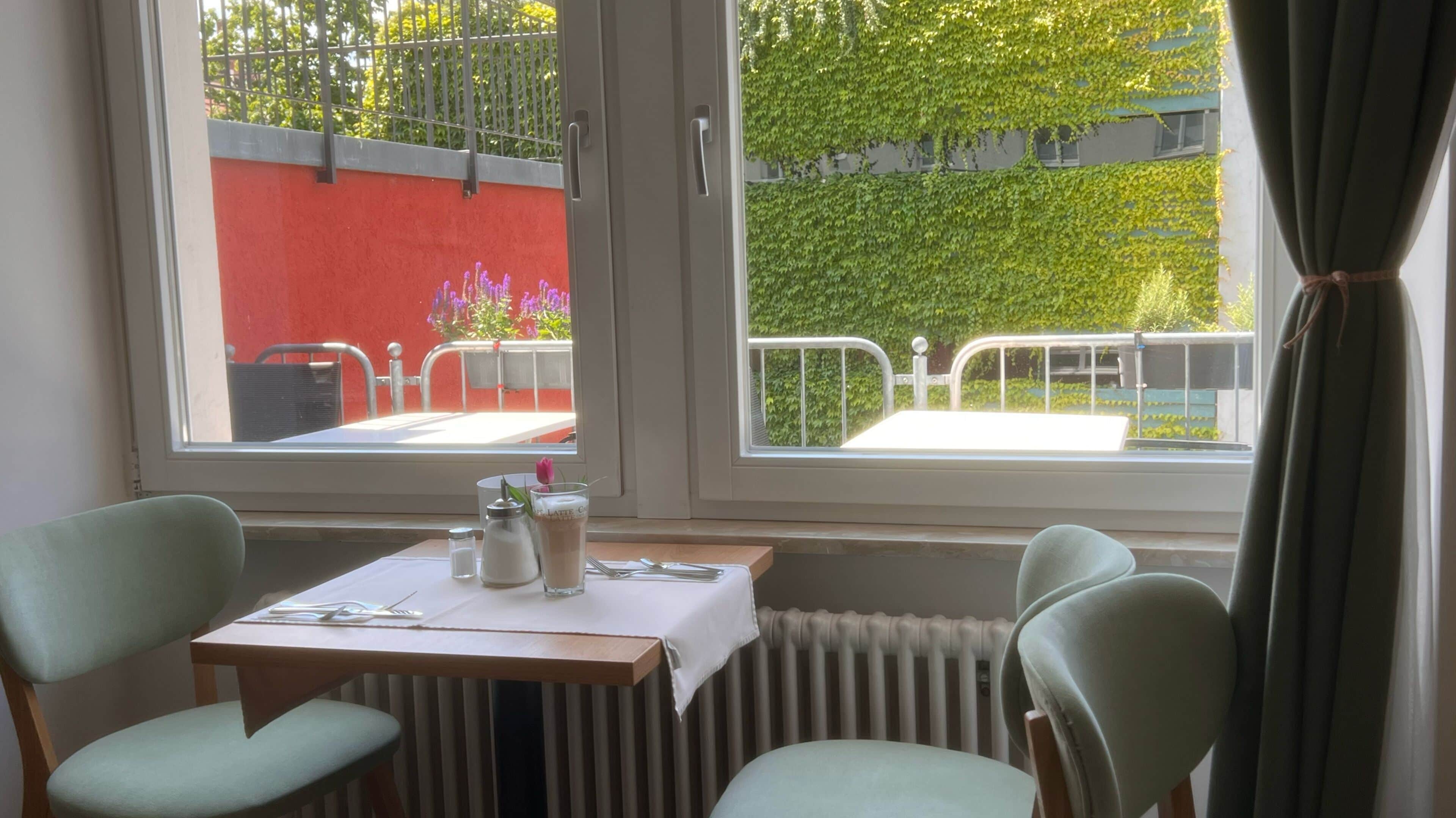 Un comedor con una mesa dispuesta para dos personas, una ventana que ofrece vistas a un muro rojo y follaje, y un radiador debajo de la ventana.