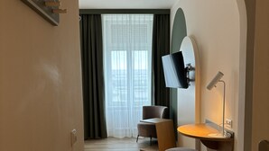 A modern hotel room with a wooden floor, a desk with a chair, a round ottoman, a flat-screen TV mounted on the wall, and a large window with curtains. - Gästehaus Centro (Konstanz)