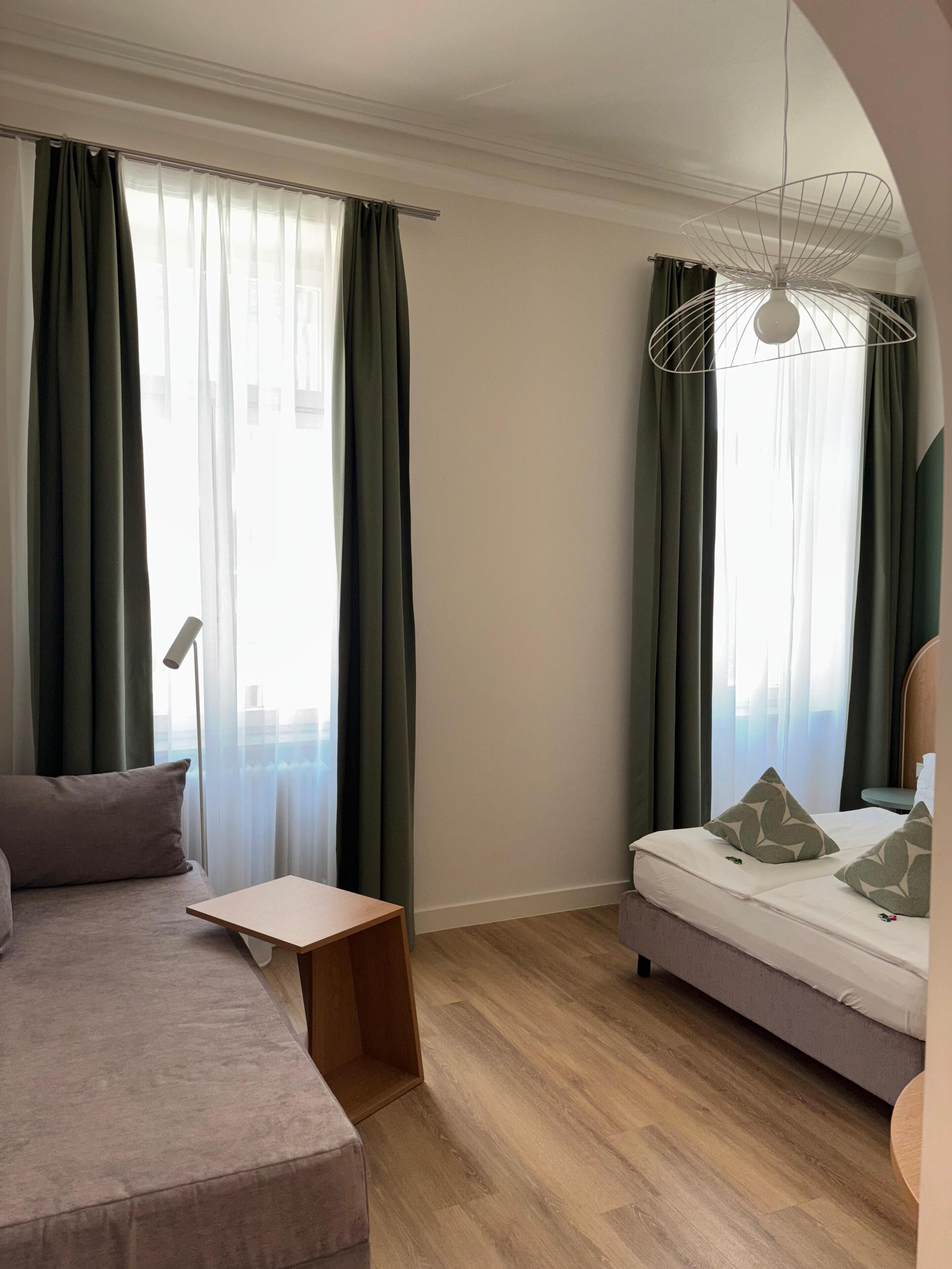 Una habitación de hotel moderna con sofá, una mesita de madera, una cama con almohadas estampadas y una lámpara de techo original.
