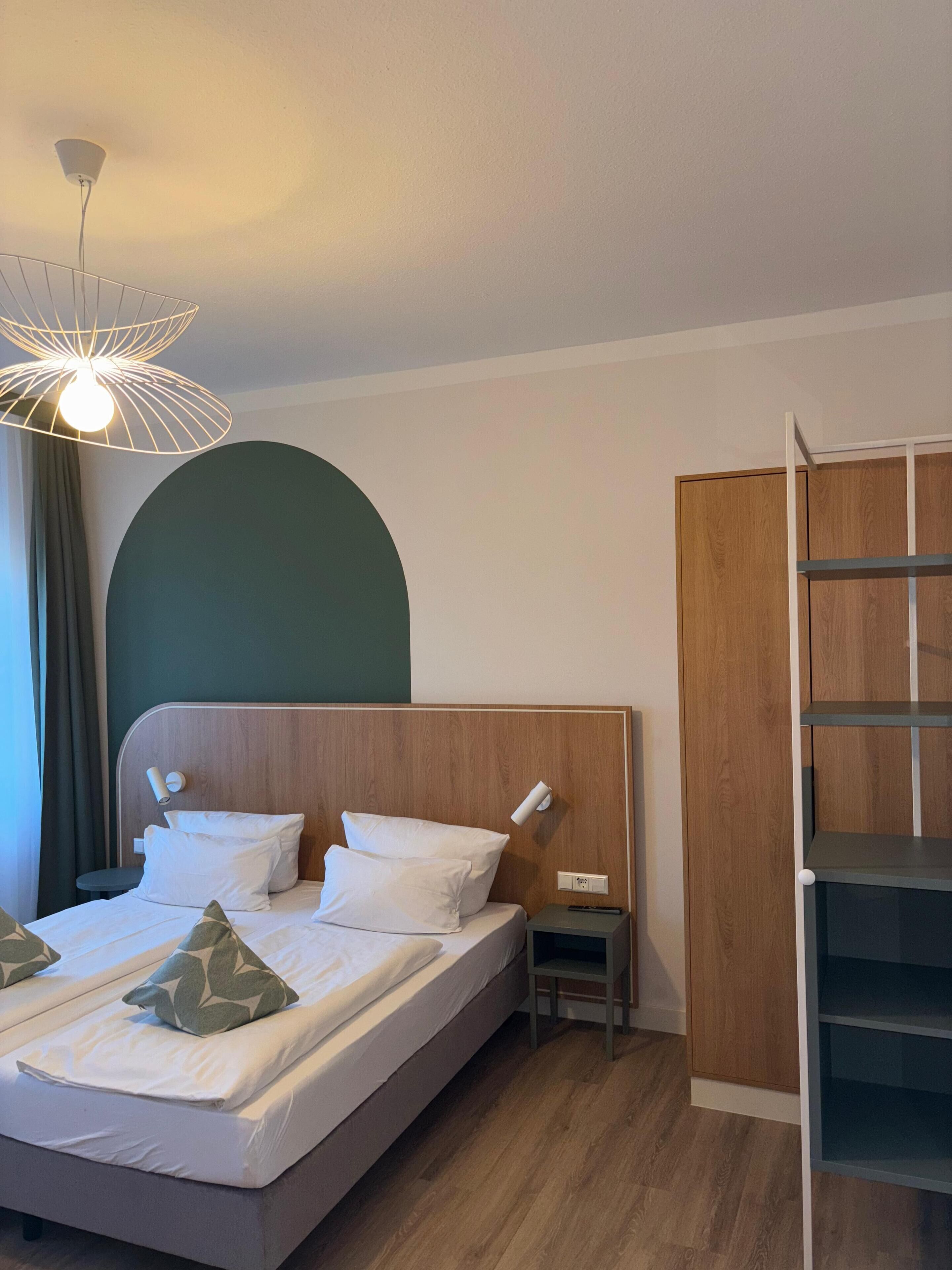 A modern hotel room with a wooden headboard, a bed with white linens, a small bedside table, a nightstand, and a built-in shelving unit.