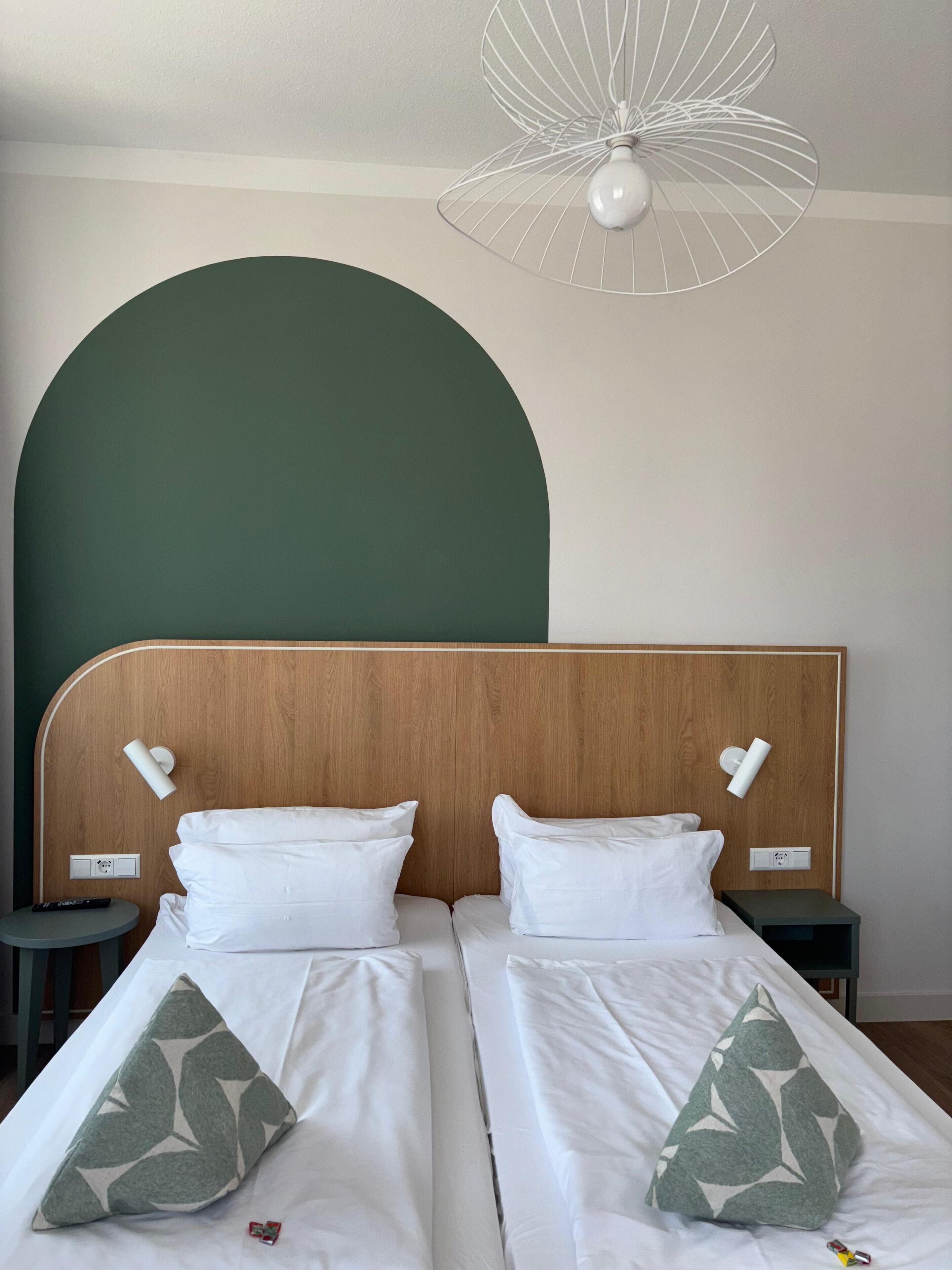A hotel room with two beds, each with white bedding and green leaf-patterned pillows. There is a wooden headboard, a white wall-mounted light fixture, and a ceiling fan.
