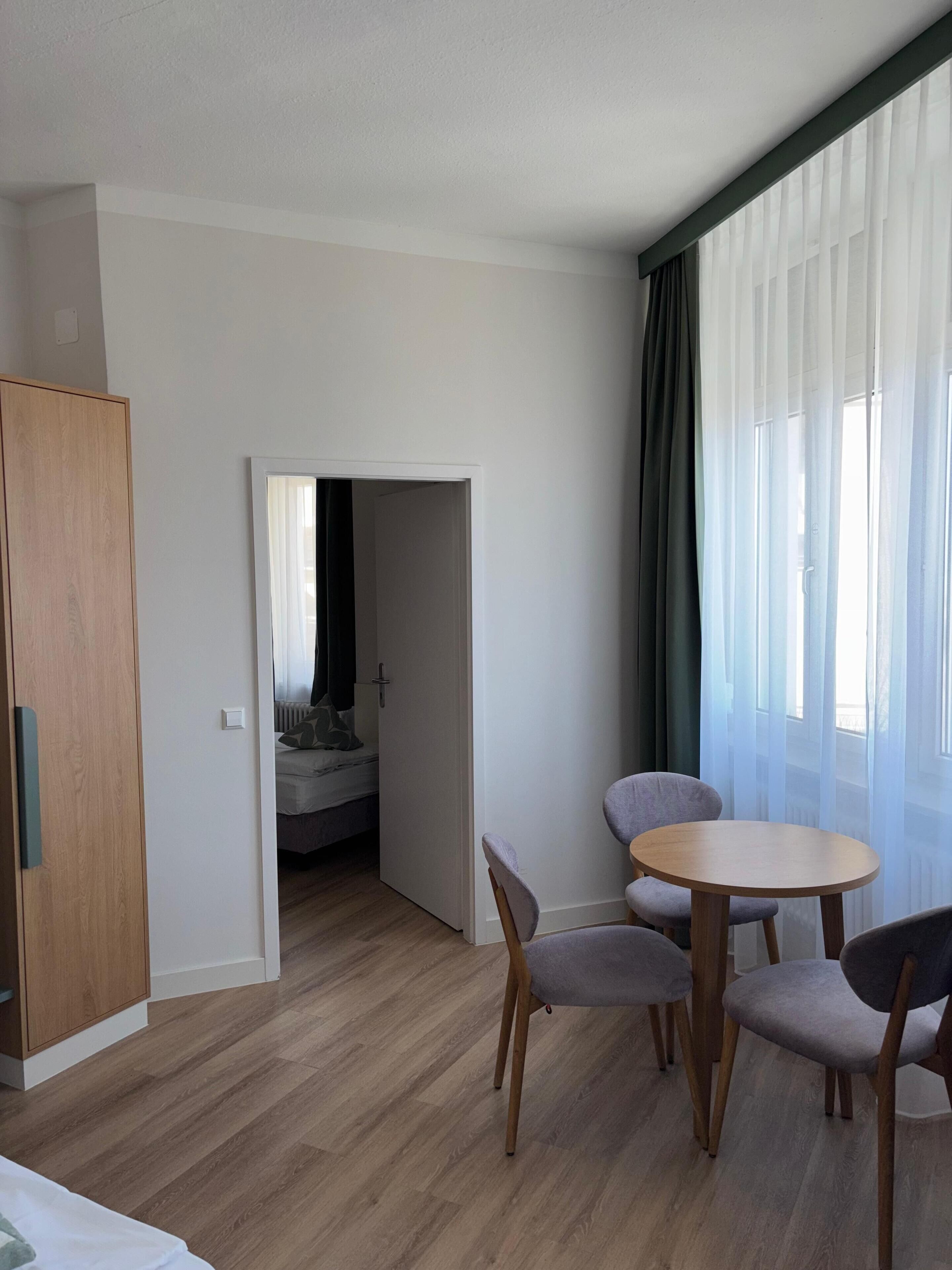 A hotel room with a wooden wardrobe, a small round table with chairs, a bed in the background, and sheer curtains on the windows.
