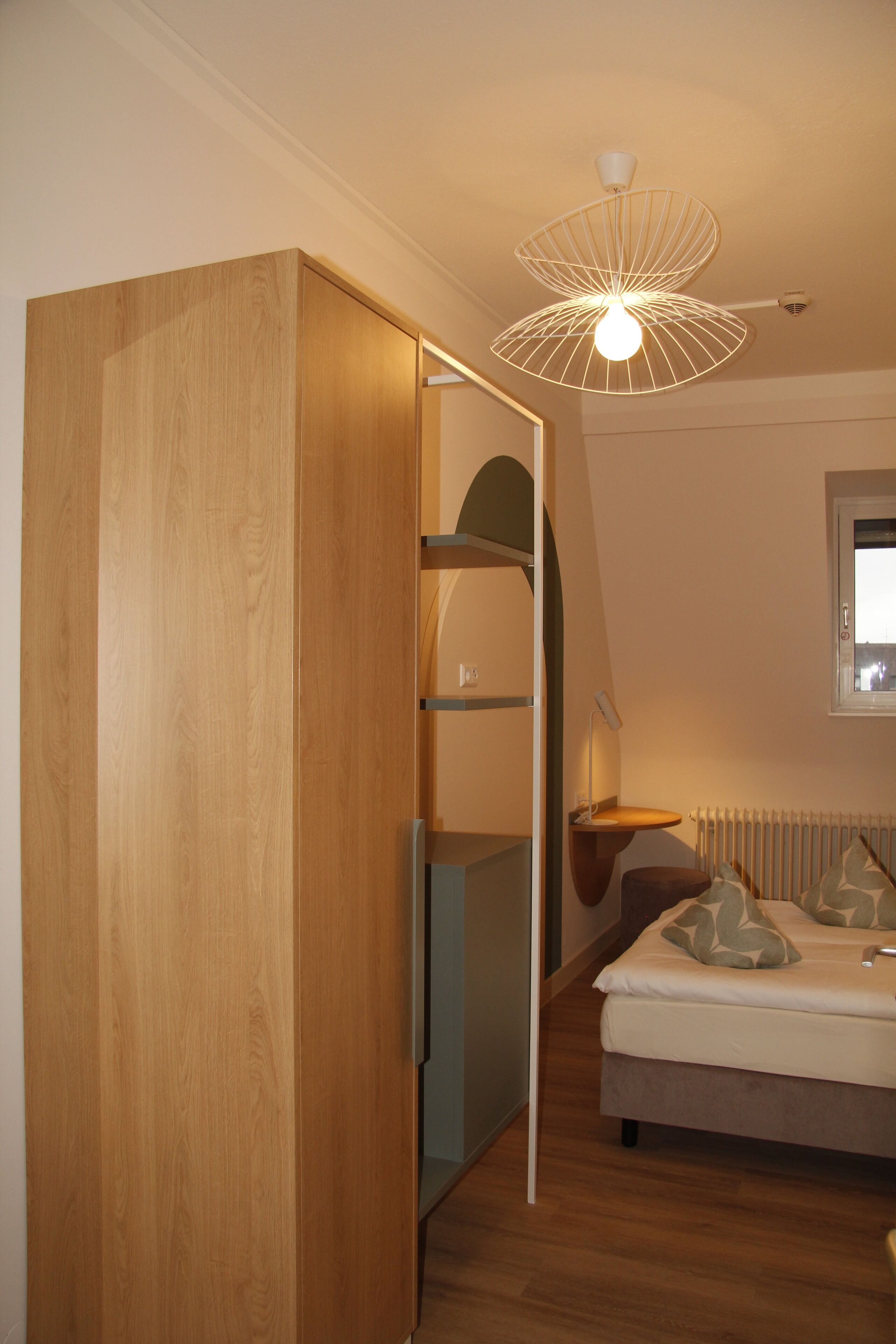 Une chambre avec une armoire en bois, un lit agrémenté d’oreillers à motifs, un radiateur et un luminaire fixé au plafond.