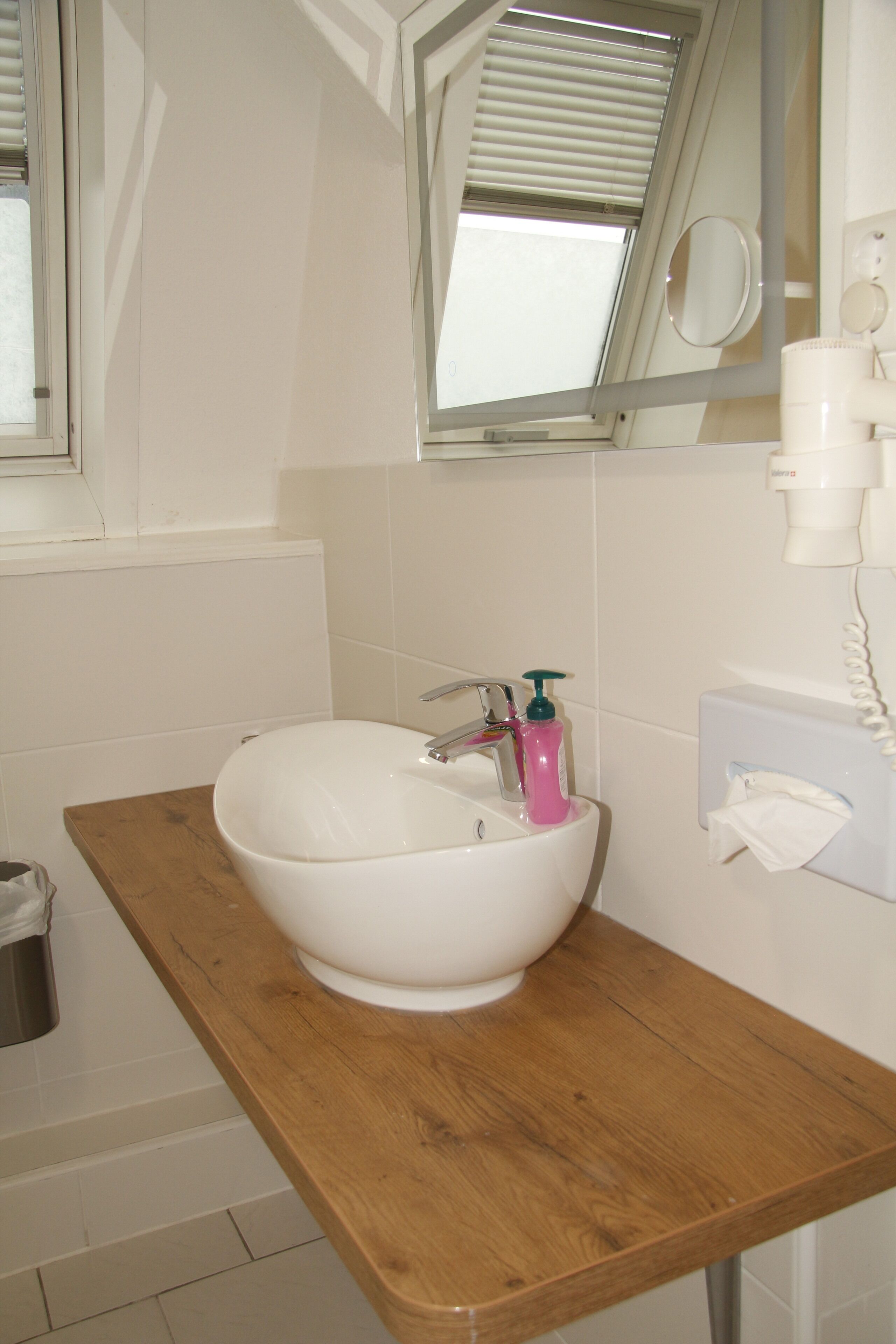 Une salle de bain avec un lavabo blanc, un plan de toilette en bois et une fenêtre avec des stores.