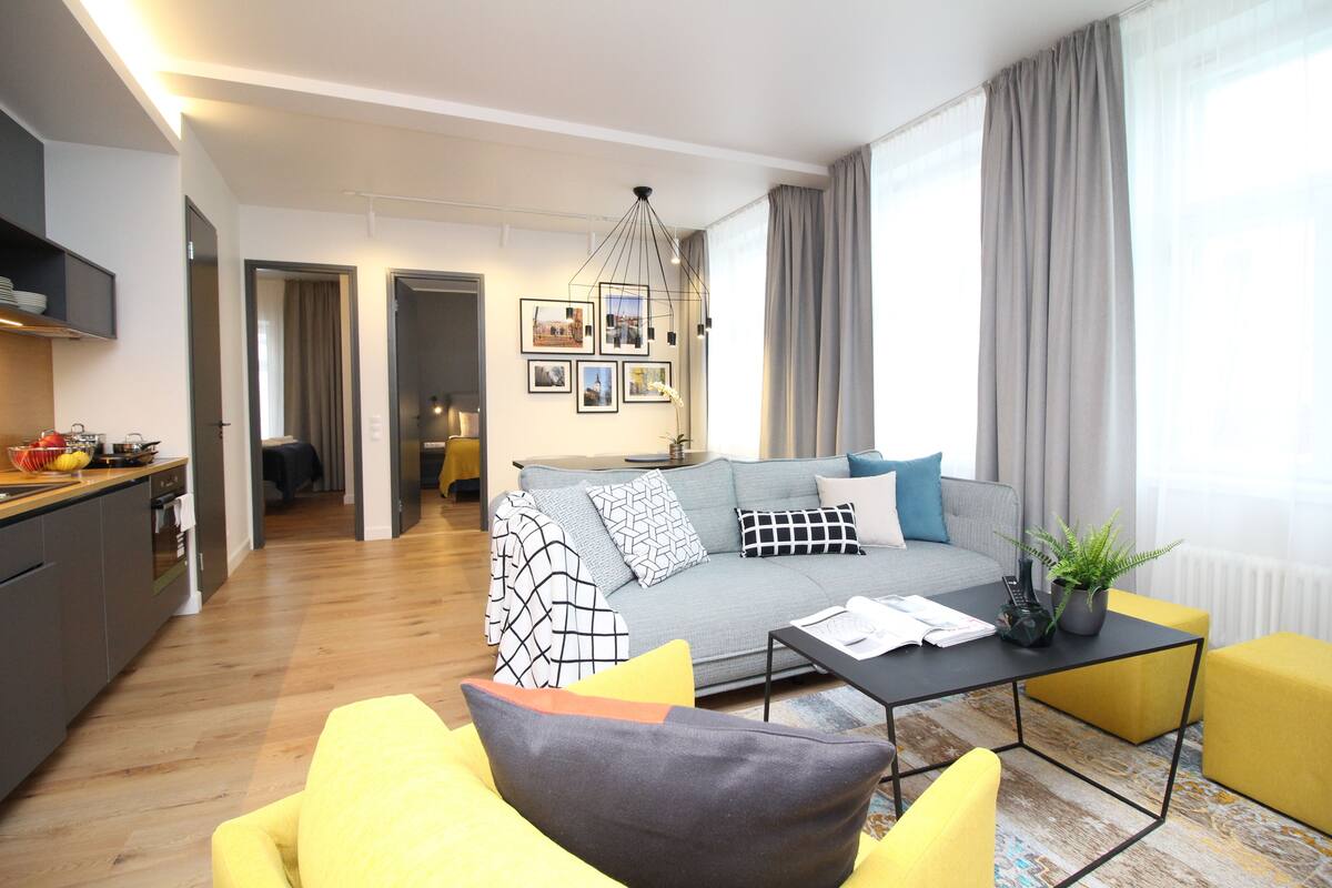 Uma sala moderna com um sofá, poltronas amarelas, uma mesa de centro e um espaço de jantar, com uma cozinha ao fundo.