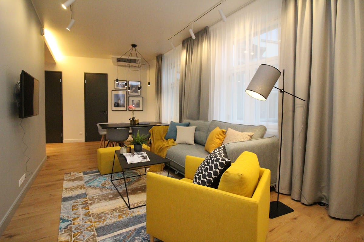 Uma sala moderna com sofá, poltrona e mesa de centro, uma área de jantar com mesa e cadeiras, e uma grande janela com cortinas.
