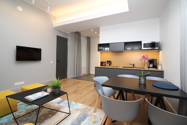 Ein modernes Wohnzimmer mit Flachbildfernseher, einem Esstisch mit Stühlen, einer Kaffeemaschine und einer Küchenzeile mit Mikrowelle.