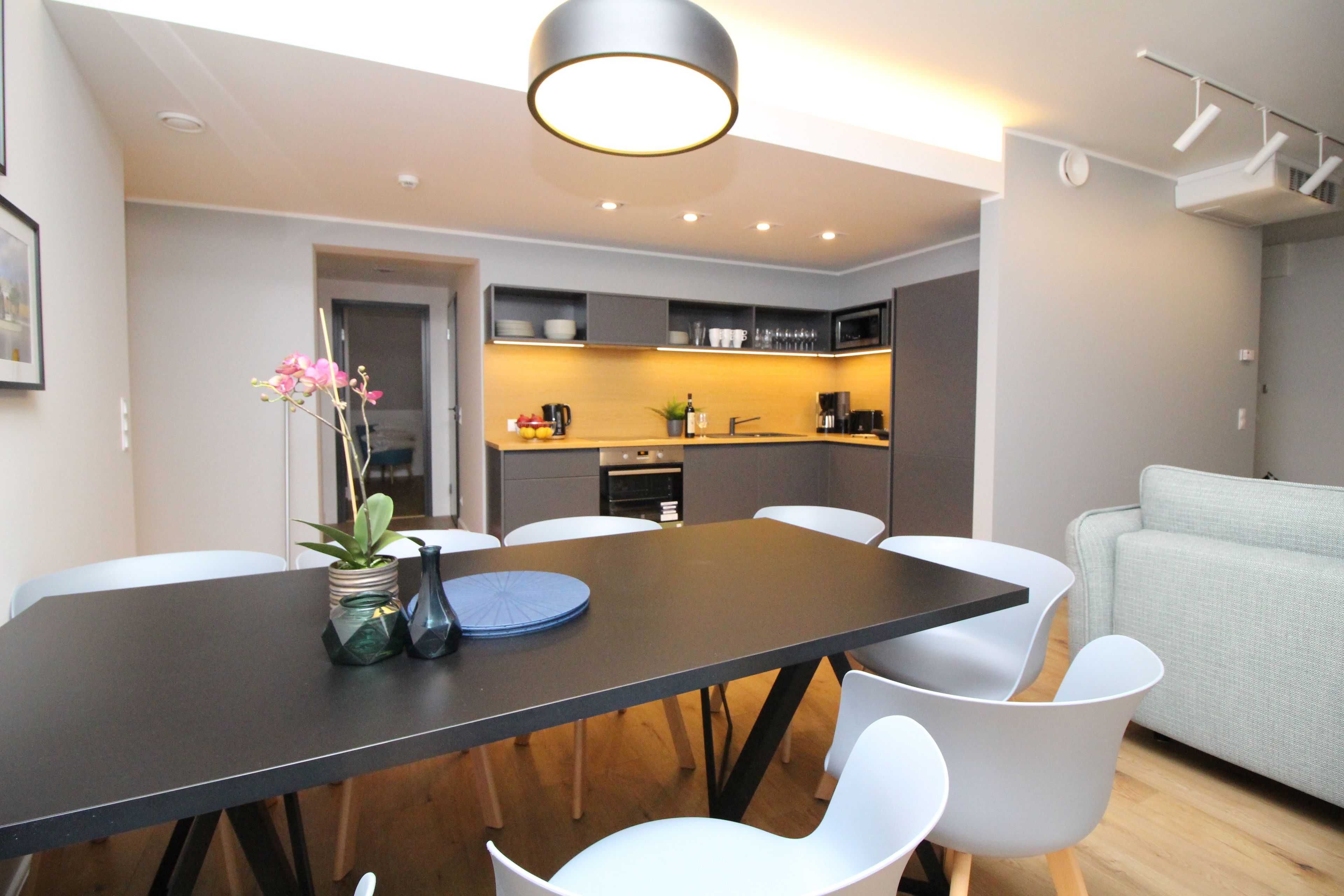 Un salon moderne comprenant une table à manger, des chaises blanches, un canapé gris et un coin cuisine avec des placards et des appareils électroménagers.