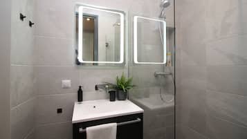 Un baño moderno con un lavamanos blanco, un espejo con iluminación perimetral, una planta en maceta y una ducha con cerramiento de vidrio.