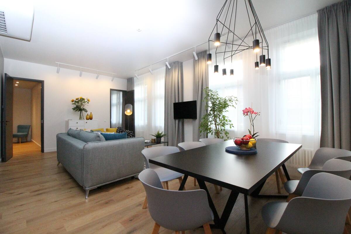 Uma sala de estar moderna com um sofá cinzento, uma mesa de jantar com cadeiras, uma televisão de tela plana e uma janela grande com cortinas.